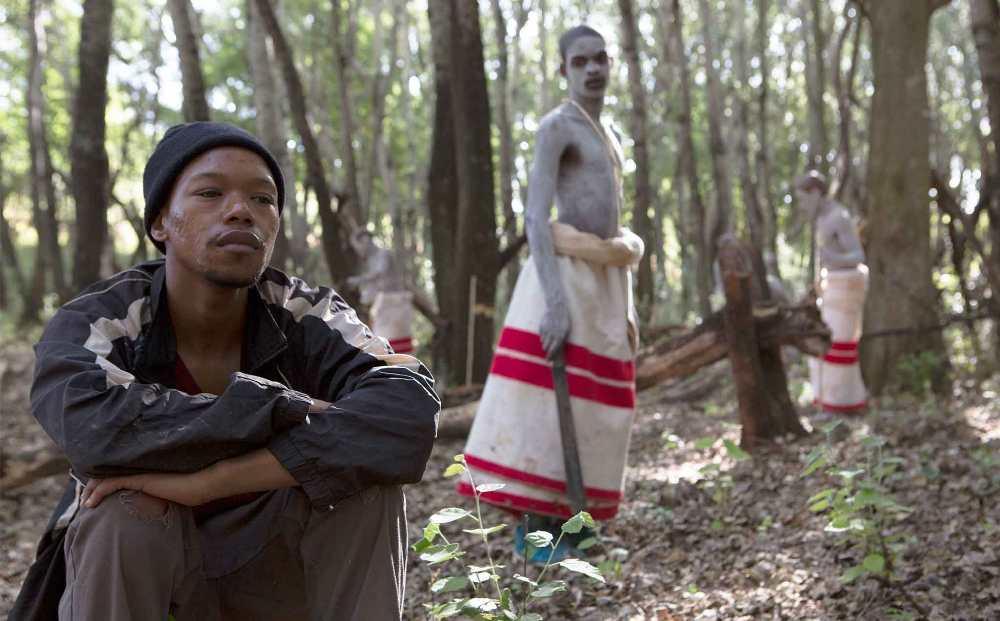 Film still from The Wound (2017): Protagonist squats in forest with figures in background