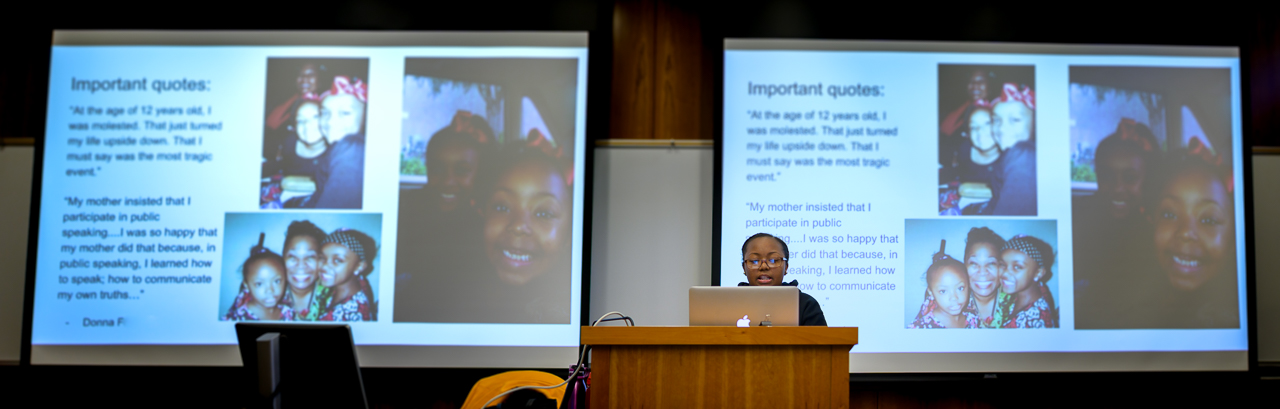Carleese B. presents at lectern in front of two large projection screens