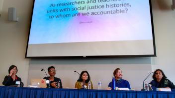 Low angle wide view of panelists beneath slide that reads, "As researchers and teachers from units with social justice histories, to whom are we accountable?"