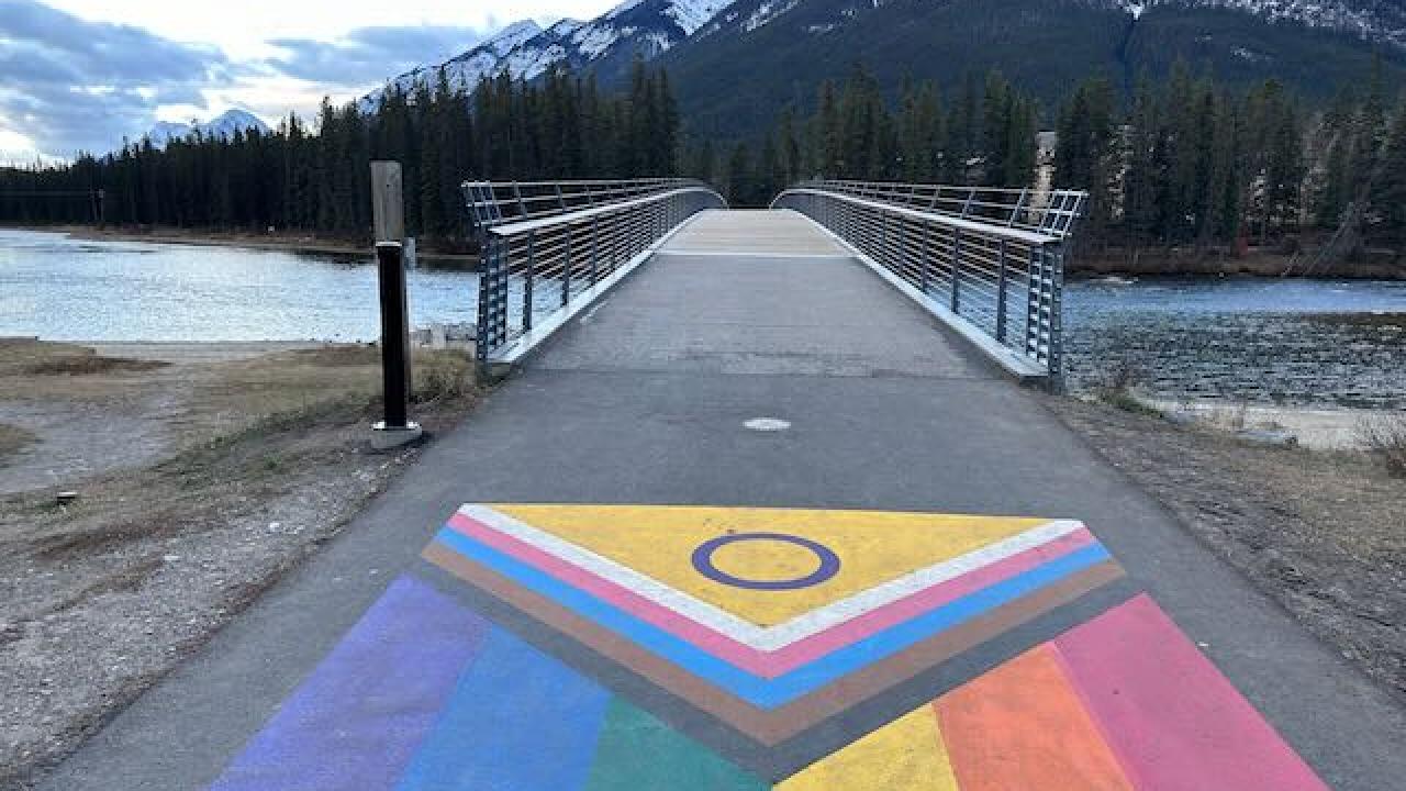 LGBTQ+ flag painted on sidewalk in front of a bridge over a river with a mountain in the background
