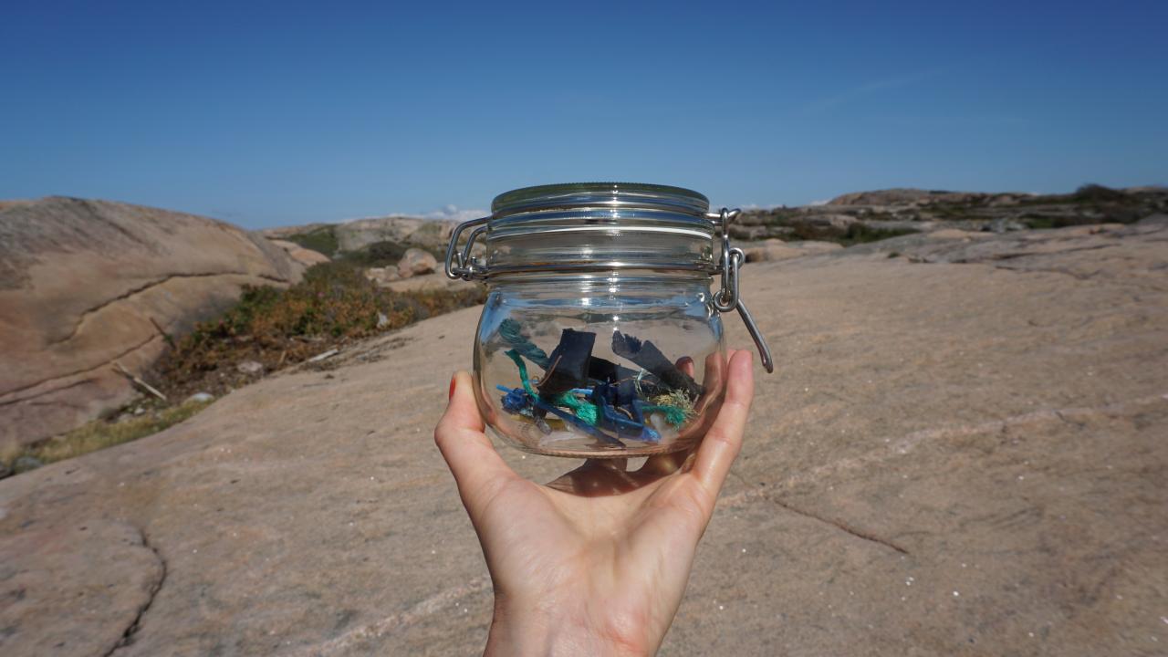 A photo of a hand holding onto a jar filled with plastic off a coast.