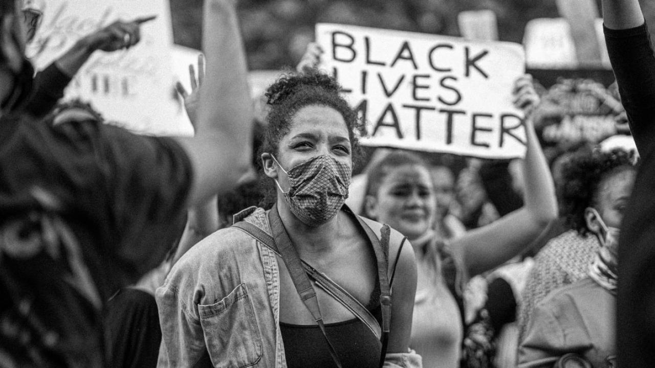 Black and white photo of a masked BLM protester with a sign behind them reading "Black Lives Matter"