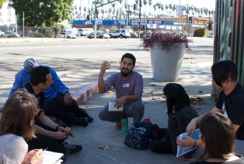 Kneeling Latinx person speaking to a circle of a half dozen people seated on the ground, in the background is a wide city street with rows of tall palm trees