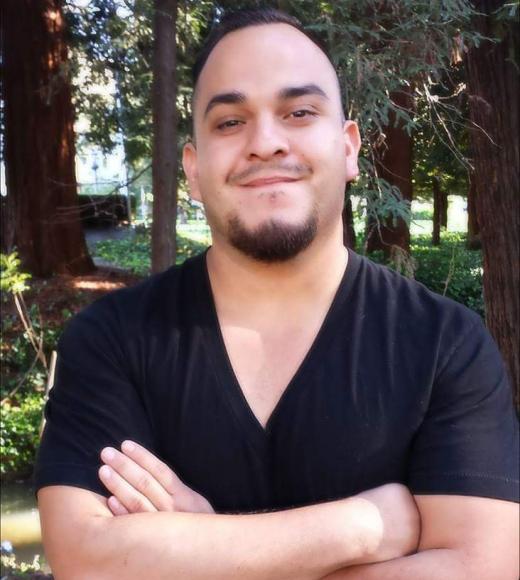 Smiling man with crossed arms in black V-neck shirt among tall trees