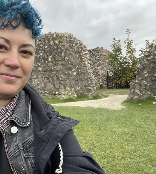 Harlin/Hayley with blue curly hair standing in front of a grassy field with stone strutctures in the background.