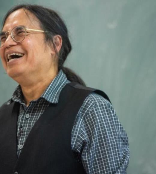Kyaw Tha, smiling, in a black vest and checkered shirt standing before a chalkboard.