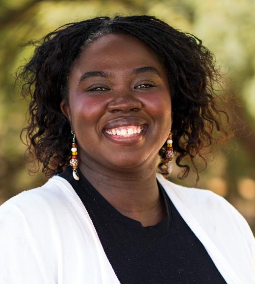 Smiling woman with black hair and beaded earrings wearing white cardigan, blurred trees