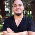 Smiling man with crossed arms in black V-neck shirt among tall trees