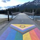 LGBTQ+ flag painted on sidewalk in front of a bridge over a river with a mountain in the background