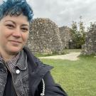 Harlin/Hayley with blue curly hair standing in front of a grassy field with stone strutctures in the background.