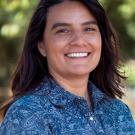 Smiling woman headshot outdoors with dark hair, blue patterned shirt, blurred green background