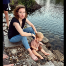 Woman with curly hair holding a toddler on rocky lakeshore, photograph