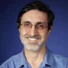 Smiling middle-aged man with glasses and gray beard in blue shirt against blue background, photo